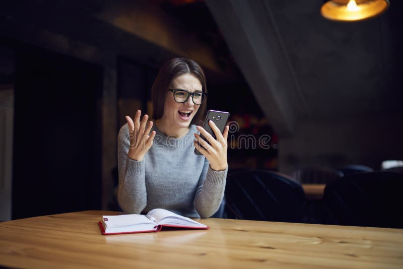 Muchacha del inconformista en lentes que lee noticias del choque en el teléfono móvil imagen de archivo
