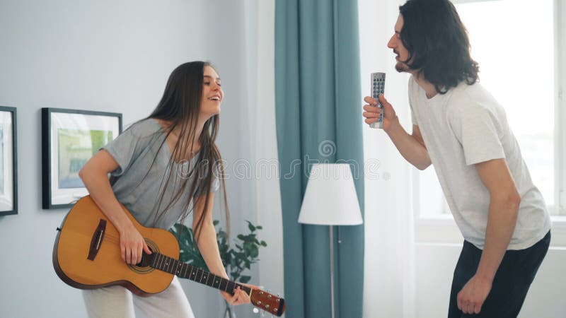 Muchacha e individuo que cantan tocando la guitarra y bailando en cama en el apartamento