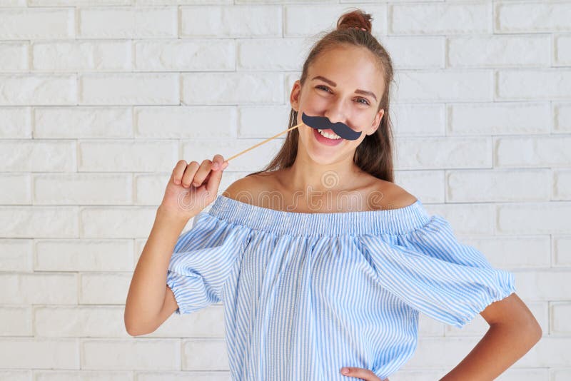 Muchacha elegante joven alegre que presenta con un bigote de papel en el sti fotos de archivo libres de regalías Muchacha elegante joven alegre que presenta con un bigote de papel en el sti fotos de archivo libres de regalías