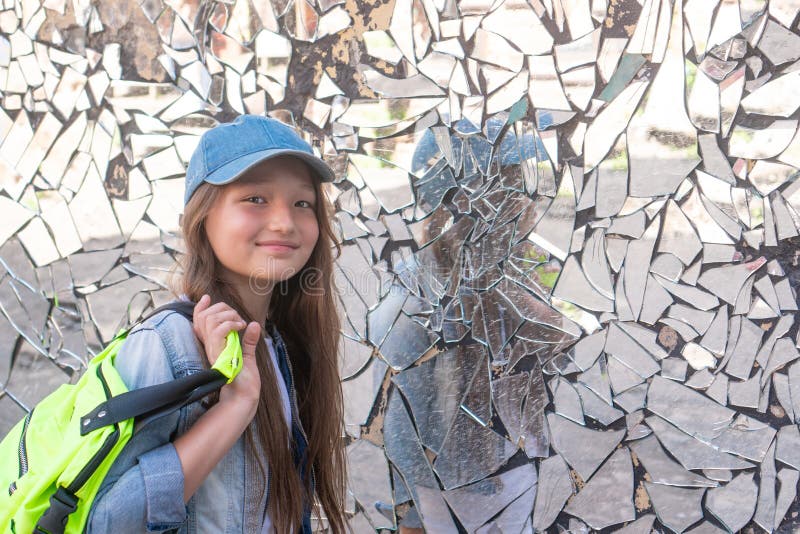 Young girl on the background of a broken mirror and smiling. Girl schoolgirl in a cap and with a backpack. Young girl on the background of a broken mirror and smiling. Girl schoolgirl in a cap and with a backpack.