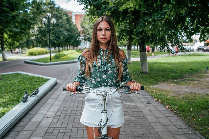Muchacha en el verano en soportes del parque con una bicicleta en la ciudad Árboles de la calzada del fondo Forma de vida activa fotos de archivo libres de regalías Muchacha en el verano en soportes del parque con una bicicleta en la ciudad Árboles de la calzada del fondo Forma de vida activa fotos de archivo libres de regalías
