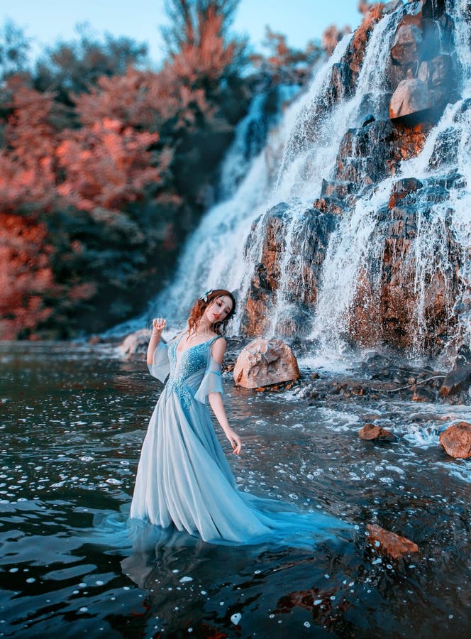 Lovely girl with fair tender skin stands in aqua in long light blue dress, mystical mermaid near high waterfall with powerful streams of cold water, Lost doll with Korean face and waterproof makeup. Lovely girl with fair tender skin stands in aqua in long light blue dress, mystical mermaid near high waterfall with powerful streams of cold water, Lost doll with Korean face and waterproof makeup.