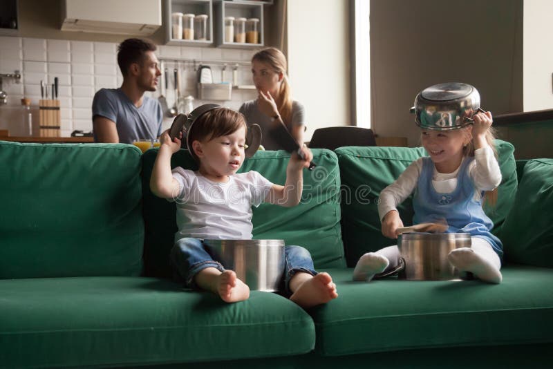 Happy toddler girl and boy playing with kitchenware on sofa at home, little sister and brother laughing using pots as drums and hats, holding kitchen utensils, making noises, spending together. Happy toddler girl and boy playing with kitchenware on sofa at home, little sister and brother laughing using pots as drums and hats, holding kitchen utensils, making noises, spending together