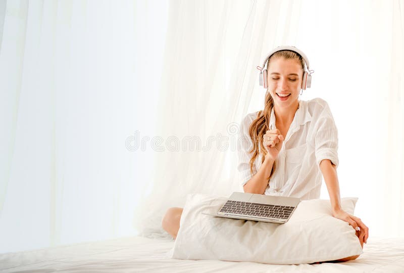 Muchacha hermosa con la estancia blanca de la camisa en cama con el ordenador portátil y escuchar la música con la emoción feliz imagen de archivo Muchacha hermosa con la estancia blanca de la camisa en cama con el ordenador portátil y escuchar la música con la emoción feliz imagen de archivo