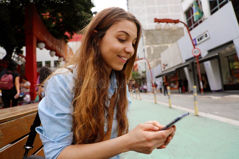 Muchacha hermosa del estudiante que se sienta en mensajería del banco de la calle con el teléfono móvil en el sao Paulo City, el imagen de archivo Muchacha hermosa del estudiante que se sienta en mensajería del banco de la calle con el teléfono móvil en el sao Paulo City, el imagen de archivo