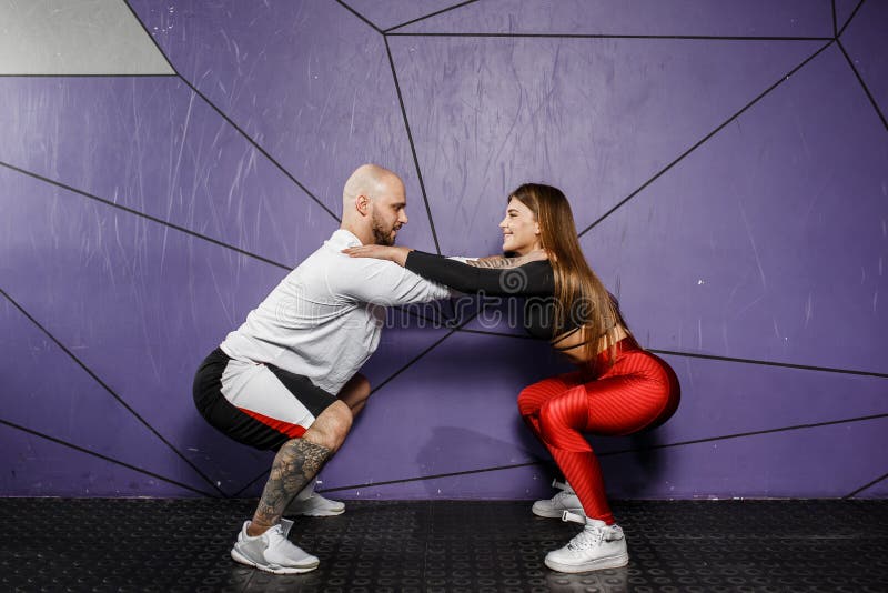 Muchacha hermosa delgada en ropa elegante de los deportes y hombre atlético fuerte hacer posiciones en cuclillas en el fondo de l foto de archivo