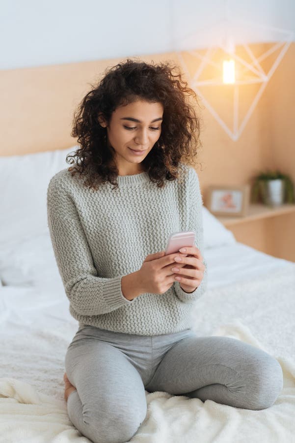 Muchacha hermosa que usa smartphone mientras que se sienta en cama imagenes de archivo Muchacha hermosa que usa smartphone mientras que se sienta en cama imagenes de archivo