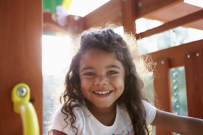 Young Hispanic girl playing on a climbing frame in a playground smiling to camera, backlit, close up. Young Hispanic girl playing on a climbing frame in a playground smiling to camera, backlit, close up