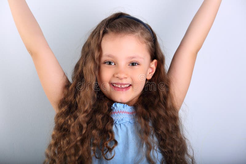 Beautiful smiling natural joying kid girl looking with long curly hair style and rises her hands up on blue background. Closeup portrait. Beautiful smiling natural joying kid girl looking with long curly hair style and rises her hands up on blue background. Closeup portrait