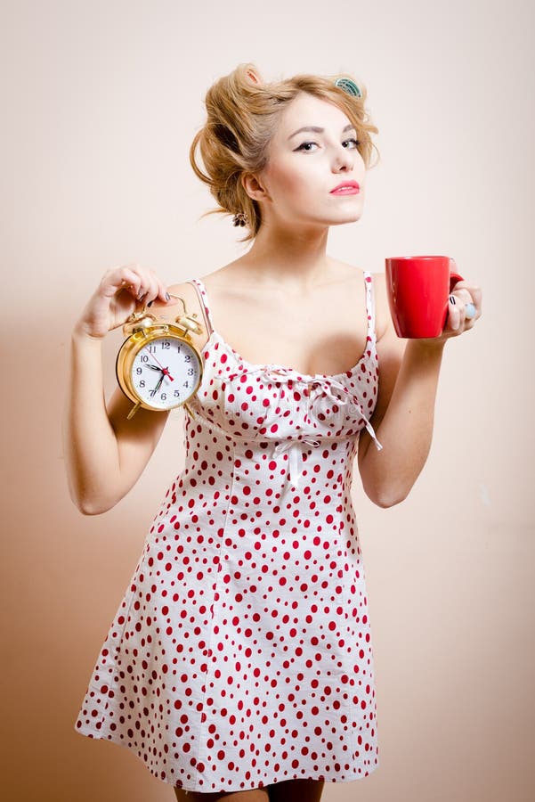 Muchacha modela divertida rubia hermosa con los bigudíes que sostienen el despertador y la taza de oro de bebida caliente que mir imagen de archivo Muchacha modela divertida rubia hermosa con los bigudíes que sostienen el despertador y la taza de oro de bebida caliente que mir imagen de archivo