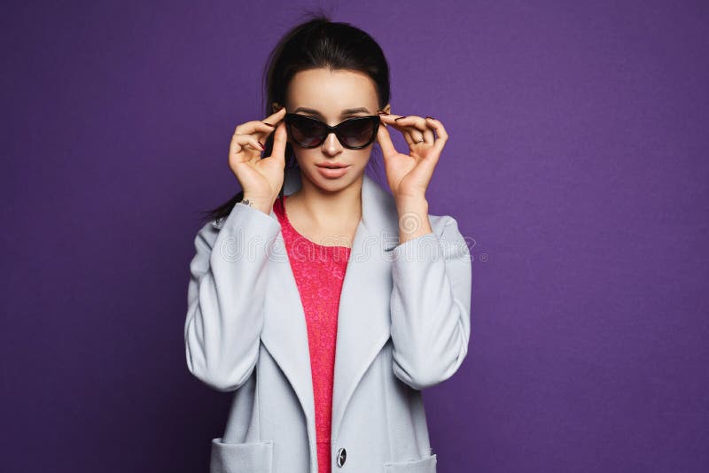 Muchacha modelo morena hermosa y de moda en vestido rojo y la capa que ajustan sus gafas de sol y que presentan en el fondo viole imagenes de archivo Muchacha modelo morena hermosa y de moda en vestido rojo y la capa que ajustan sus gafas de sol y que presentan en el fondo viole imagenes de archivo