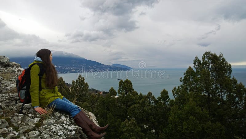 Muchacha morena con una mochila que lleva una capa y las botas que se sientan en una roca y que miran hacia el mar y las montañas imagen de archivo libre de regalías Muchacha morena con una mochila que lleva una capa y las botas que se sientan en una roca y que miran hacia el mar y las montañas imagen de archivo libre de regalías