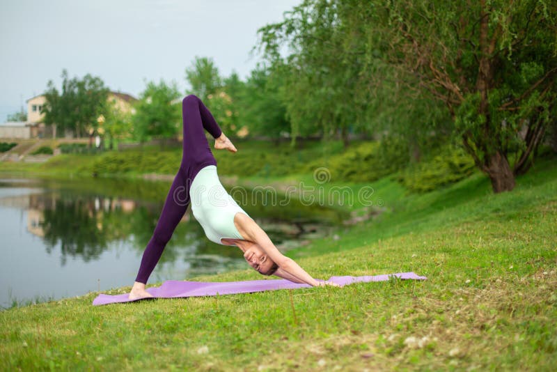 Muchacha morena delgada que hace yoga en el verano en un césped verde por el lago fotos de archivo Muchacha morena delgada que hace yoga en el verano en un césped verde por el lago fotos de archivo