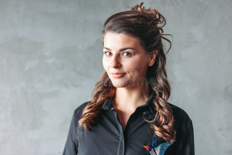 Muchacha morena sonriente encantadora joven con el pelo ondulado en vestido negro de la camisa, en fondo gris imágenes de archivo libres de regalías Muchacha morena sonriente encantadora joven con el pelo ondulado en vestido negro de la camisa, en fondo gris imágenes de archivo libres de regalías
