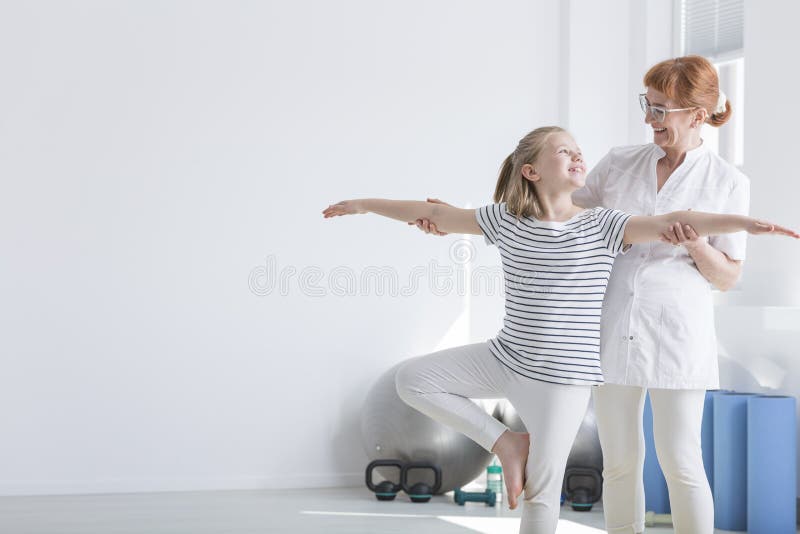 Muchacha que se coloca en una pierna. Muchacha feliz que sonríe al fisioterapeuta mientras que se coloca en una pierna durante la gimnasia correctiva