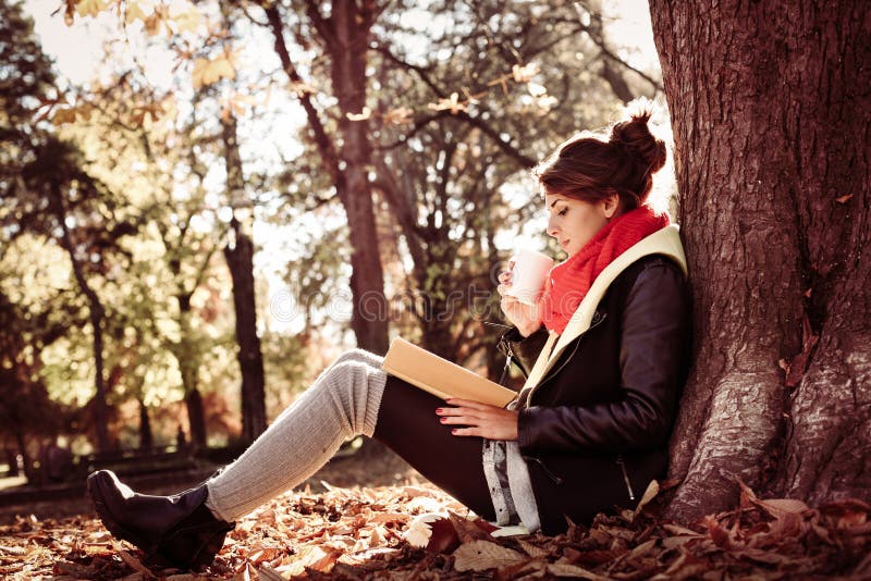 Muchacha que se inclina en un árbol en el parque y que lee el libro por mañana