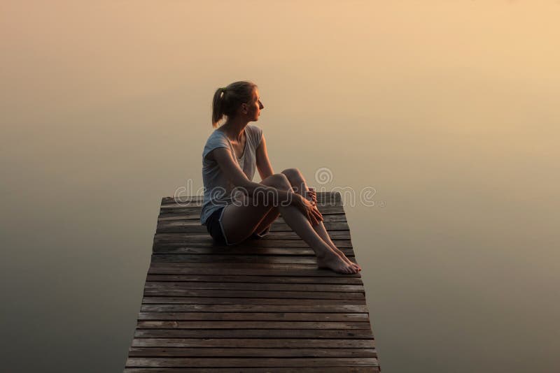 Muchacha que se sienta en un embarcadero o un embarcadero de madera en el lago en la puesta del sol foto de archivo libre de regalías Muchacha que se sienta en un embarcadero o un embarcadero de madera en el lago en la puesta del sol foto de archivo libre de regalías