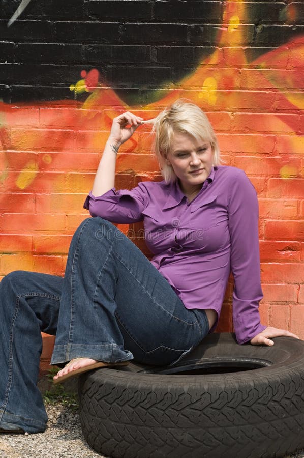 Blond woman sitting on old tire and twisting her hair. Blond woman sitting on old tire and twisting her hair.