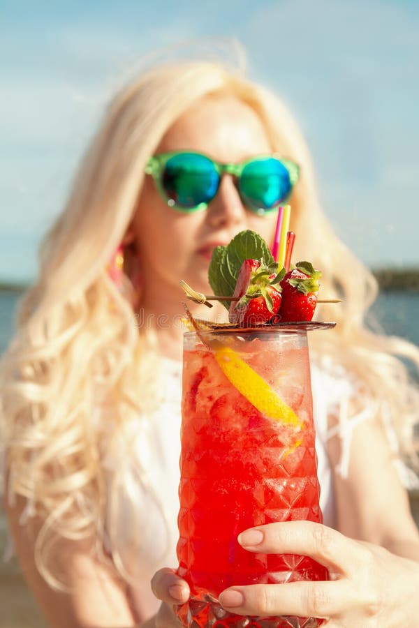 Muchacha rubia hermosa con un cóctel hermoso rojo en sus manos por el mar/el río foto de archivo libre de regalías Muchacha rubia hermosa con un cóctel hermoso rojo en sus manos por el mar/el río foto de archivo libre de regalías