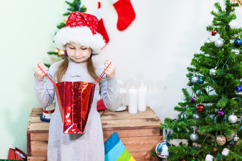 Muchacha sonriente linda en el sombrero rojo de Papá Noel que mira dentro de bolso del regalo cerca del árbol de navidad, copyspa imagen de archivo libre de regalías Muchacha sonriente linda en el sombrero rojo de Papá Noel que mira dentro de bolso del regalo cerca del árbol de navidad, copyspa imagen de archivo libre de regalías