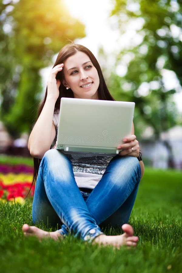 Muchacha sonriente que se sienta en hierba con la computadora portátil imagen de archivo libre de regalías Muchacha sonriente que se sienta en hierba con la computadora portátil imagen de archivo libre de regalías