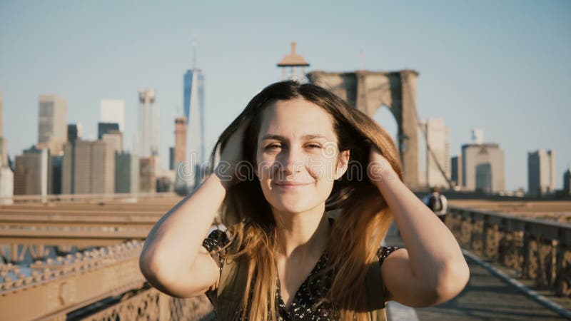 Muchacha turística europea joven con la mochila que presenta en la cámara, pelo conmovedor y sonriendo en el puente de Brooklyn