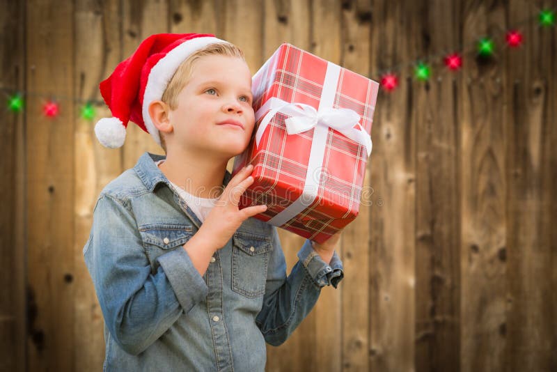 Muchacho curioso que lleva a Santa Hat Holding Christmas Gift en la madera fotos de archivo libres de regalías Muchacho curioso que lleva a Santa Hat Holding Christmas Gift en la madera fotos de archivo libres de regalías