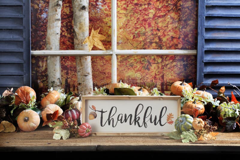 The word Thankful on white board with wood frame sitting on old wood tabletop surrounded with fall gourds and leaves with view out window to birch tree trunks and trees with fall foliage. The word Thankful on white board with wood frame sitting on old wood tabletop surrounded with fall gourds and leaves with view out window to birch tree trunks and trees with fall foliage.