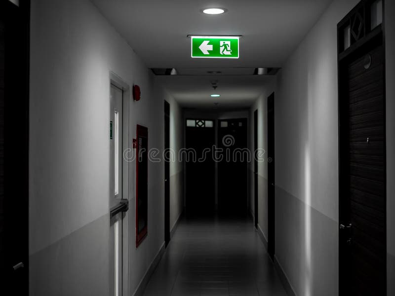 Muestra verde del fuego de la salida que cuelga en techo en el pasillo misterioso oscuro en el edificio cerca de puerta de la sal fotografía de archivo Muestra verde del fuego de la salida que cuelga en techo en el pasillo misterioso oscuro en el edificio cerca de puerta de la sal fotografía de archivo