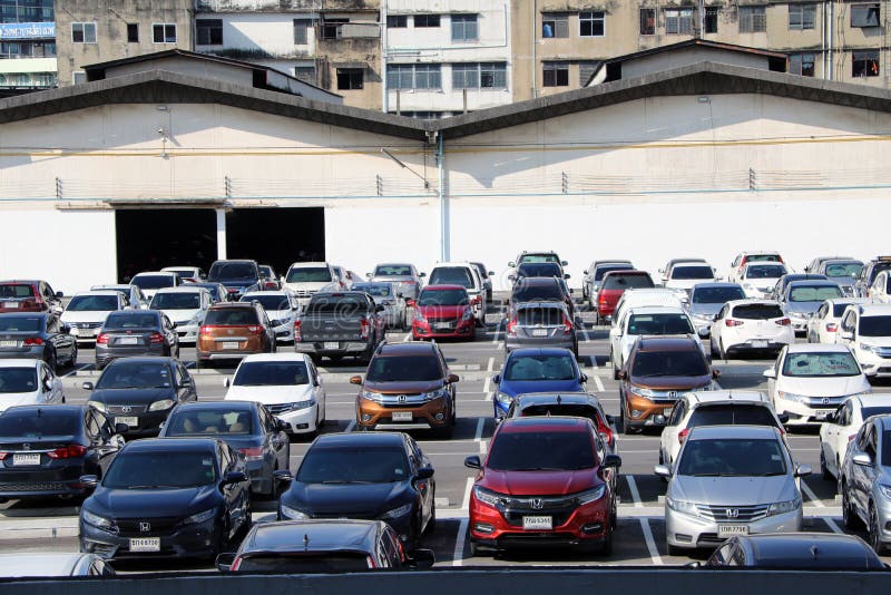 Muitos carros são estacionados no parque de estacionamento exterior com construção da garagem e construção mais alta