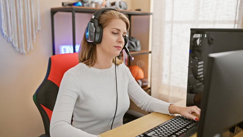 Mujer adulta enfocada con pelo rubio corto y audífonos usando una computadora en una sala de juegos en casa fotos de archivo libres de regalías