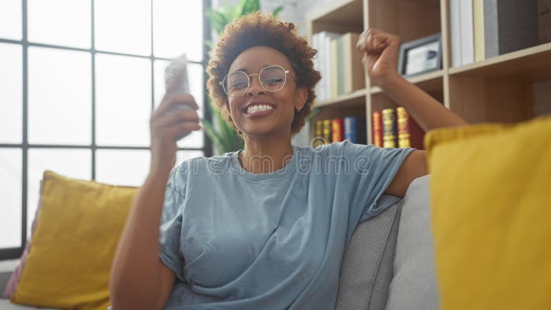 Mujer africana alegre con gafas celebrando el éxito en un acogedor salón foto de archivo libre de regalías