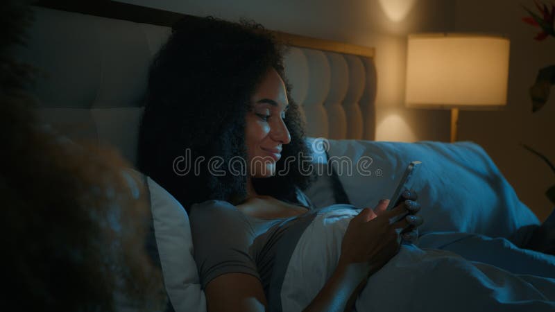 Mujer africana guapa usa smartphone tendido en la cama insomnio de noche relajándose en el dormitorio del hogar teléfono móvil fotografía de archivo