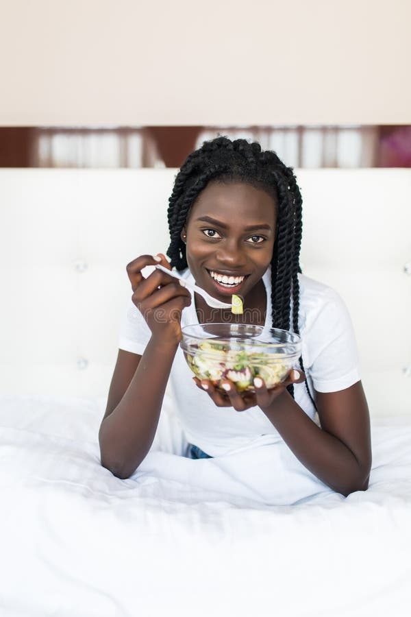 Mujer africana sana joven que se sienta en cama y que come la ensalada foto de archivo libre de regalías Mujer africana sana joven que se sienta en cama y que come la ensalada foto de archivo libre de regalías