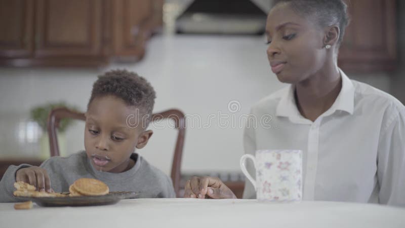 Mujer afroamericana atractiva del retrato que se sienta con su peque?o hijo por la tabla en la cocina Mam? de la relaci?n y metrajes Mujer afroamericana atractiva del retrato que se sienta con su peque?o hijo por la tabla en la cocina Mam? de la relaci?n y metrajes