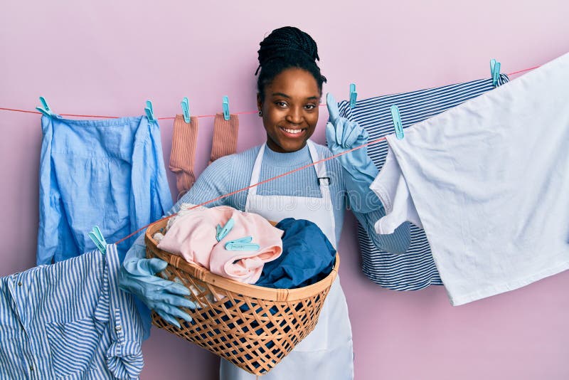 Mujer afroamericana con el pelo trenzado lavando la ropa con una cesta de mimbre sonriendo con una idea o pregunta apuntando con e. Mujer afroamericana con pelo trenzado lavando la ropa con una cesta de mimbre sonriendo con una idea o una pregunta apuntando con el dedo contenta número uno