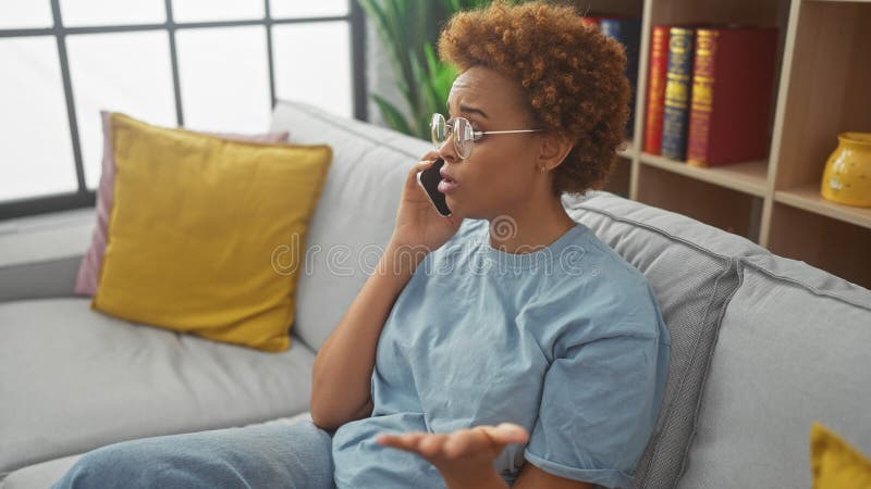 Mujer afroamericana con gafas hablando por teléfono mientras está sentada en un sofá interior imagenes de archivo