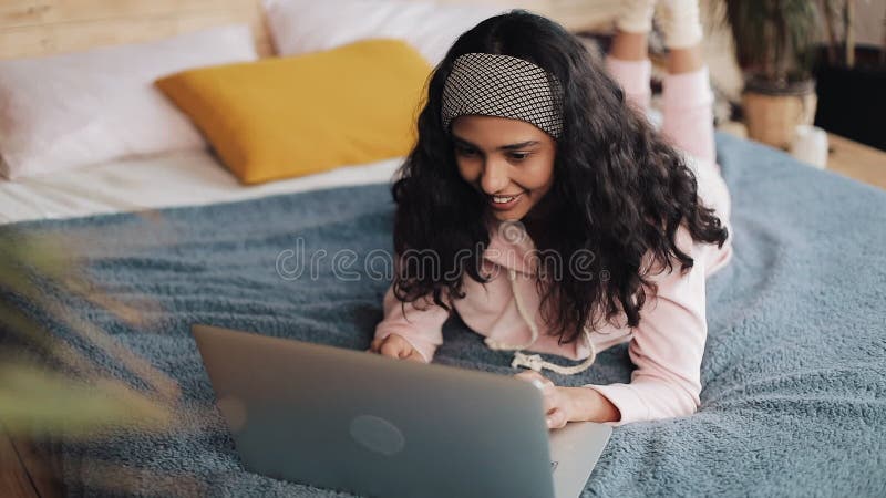 Mujer afroamericana feliz que miente en cama usando el ordenador portátil para hojear la web Muchacha que lleva la sonrisa rosada