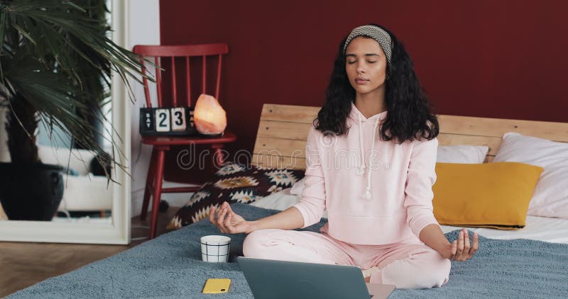 Mujer afroamericana joven hermosa que hace ejercicio de la yoga y que mira la lección preceptoral en el ordenador portátil en cas