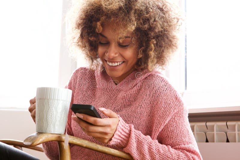 Mujer afroamericana joven que bebe la taza de café caliente y que mira el teléfono móvil imagenes de archivo