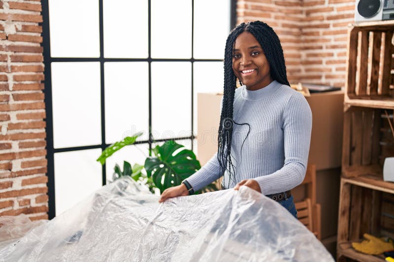 Mujer afroamericana sonriendo confiado cubriendo el sofá con plástico en su nueva casa imagen de archivo