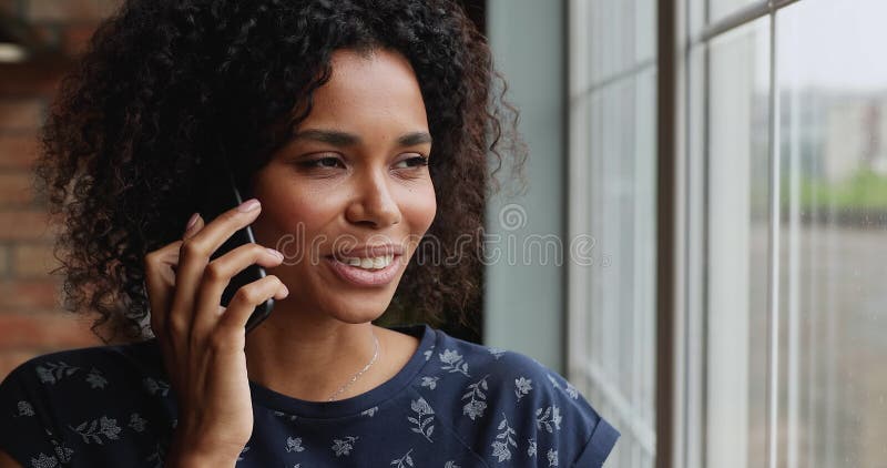 Mujer afroamericana sonriente parada al lado de la ventana hablando por teléfono