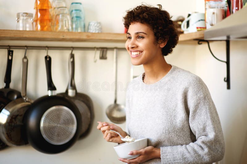 Mujer afroamericana sonriente parada en la cocina comiendo cereales para el desayuno fotos de archivo libres de regalías Mujer afroamericana sonriente parada en la cocina comiendo cereales para el desayuno fotos de archivo libres de regalías