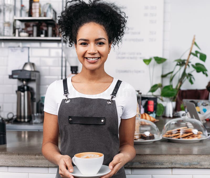 Mujer afroamericana trabajando como barista sosteniendo una taza con un capuchino recién preparado fotografía de archivo libre de regalías