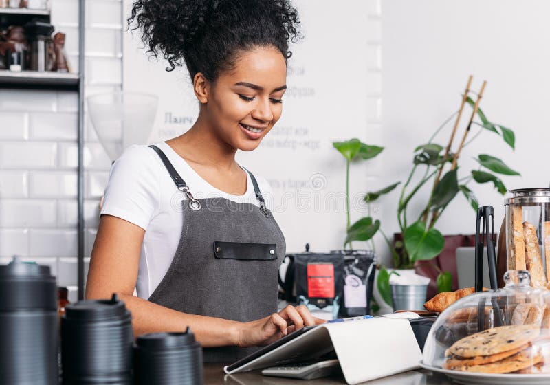 Mujer alegre con pelo rizado trabajando como barista en una cafetería usando una tableta digital foto de archivo