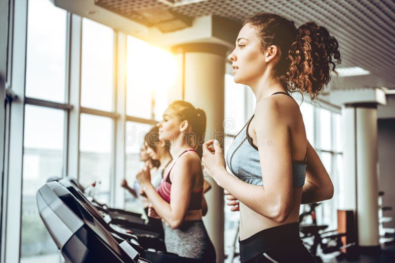 Mujer alegre joven hermosa en la ropa de deportes que corre en la rueda de ardilla en el gimnasio con otras mujeres fotos de archivo libres de regalías Mujer alegre joven hermosa en la ropa de deportes que corre en la rueda de ardilla en el gimnasio con otras mujeres fotos de archivo libres de regalías
