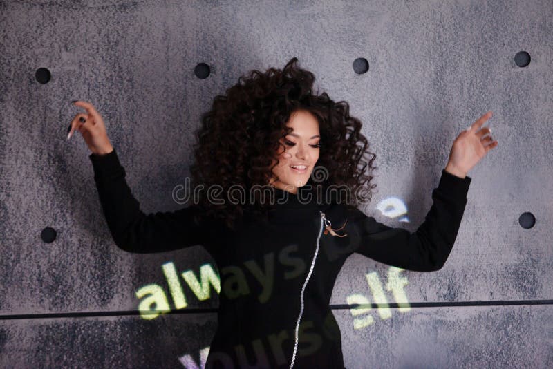 Mujer alegre y rizada bailando sobre un fondo de pared de concreto. InscripciÃ³n coloreada e iluminada dice: 'SÃ© siempre tÃº mism imagen de archivo libre de regalías