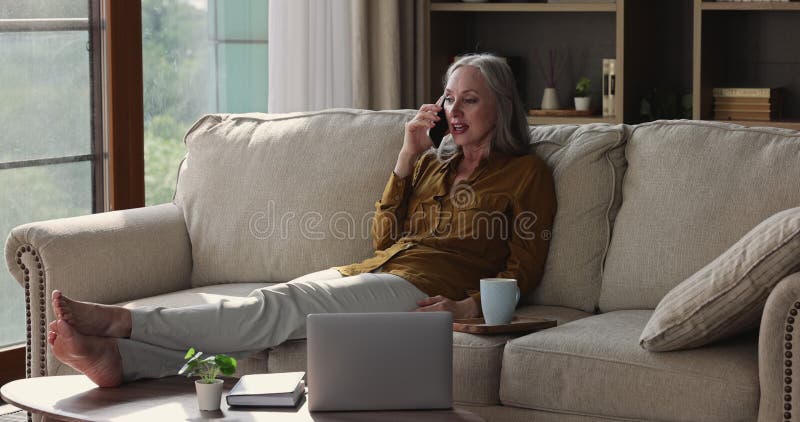 Mujer anciana se relaja en el sofá guarda el teléfono celular hacer una llamada personal