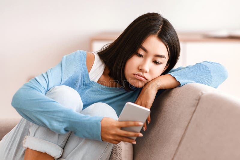 Mujer asiática frustrada mirando el teléfono móvil sentado en el sofá fotografía de archivo