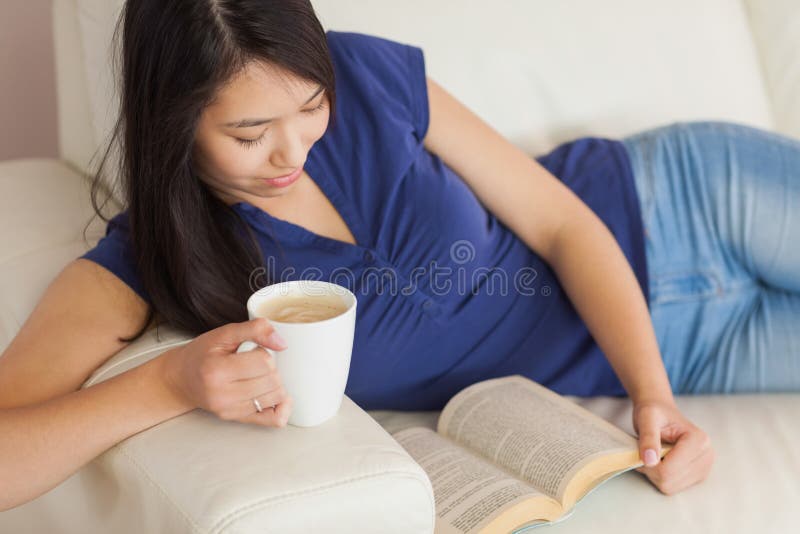 Mujer asiática joven feliz que miente en el sofá que lee una tenencia del libro foto de archivo Mujer asiática joven feliz que miente en el sofá que lee una tenencia del libro foto de archivo